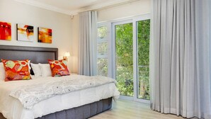 A modern bedroom with a large bed, grey curtains, and a view of greenery outside. - Stylish 2 Bedroom Apartment in Camps Bay Asilvertide (Cape Town)