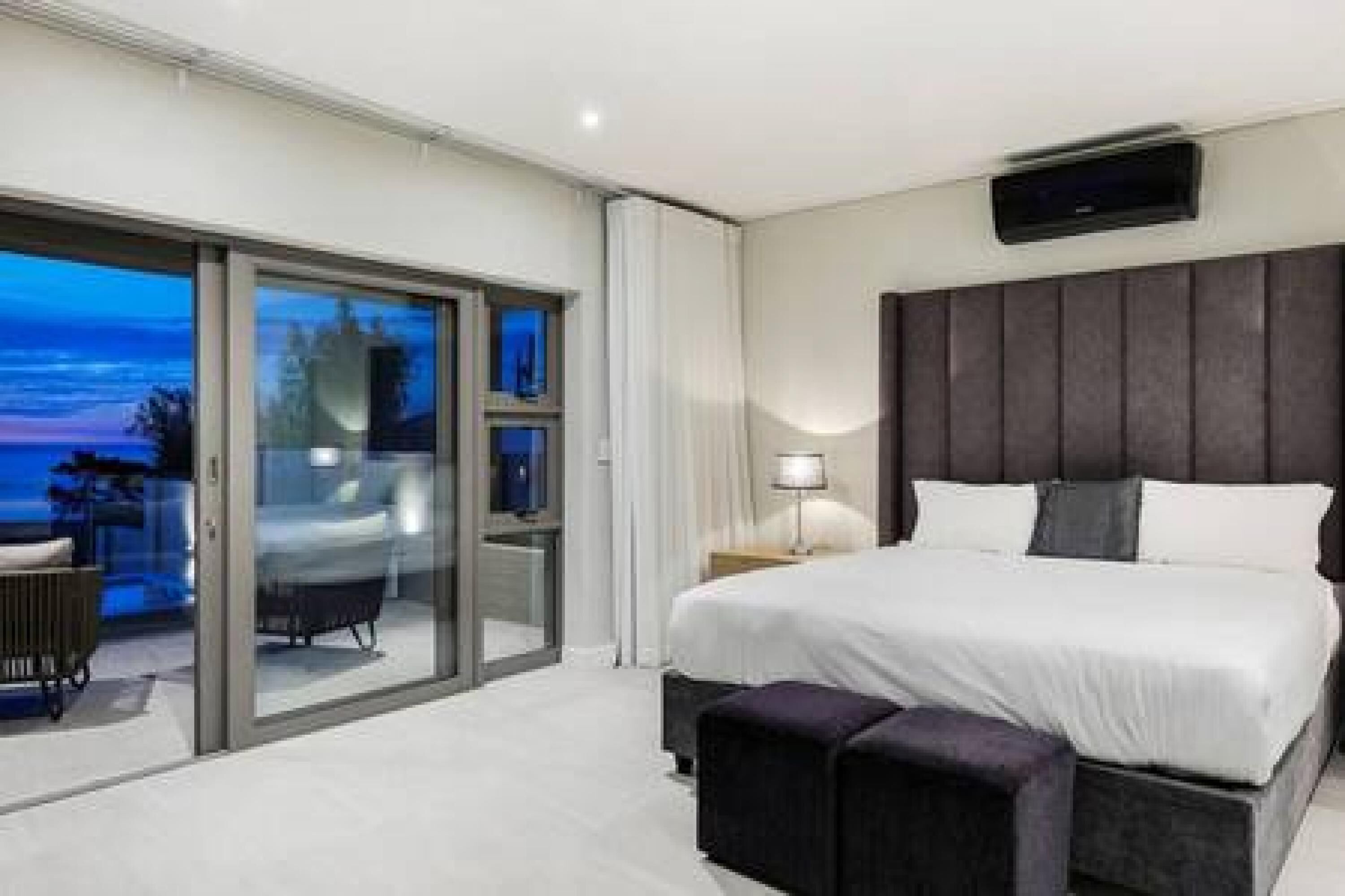 Habitación de hotel moderna con una cama grande, una mesita de noche, una lámpara y un ventanal corredero que da a un balcón con vistas al océano.
