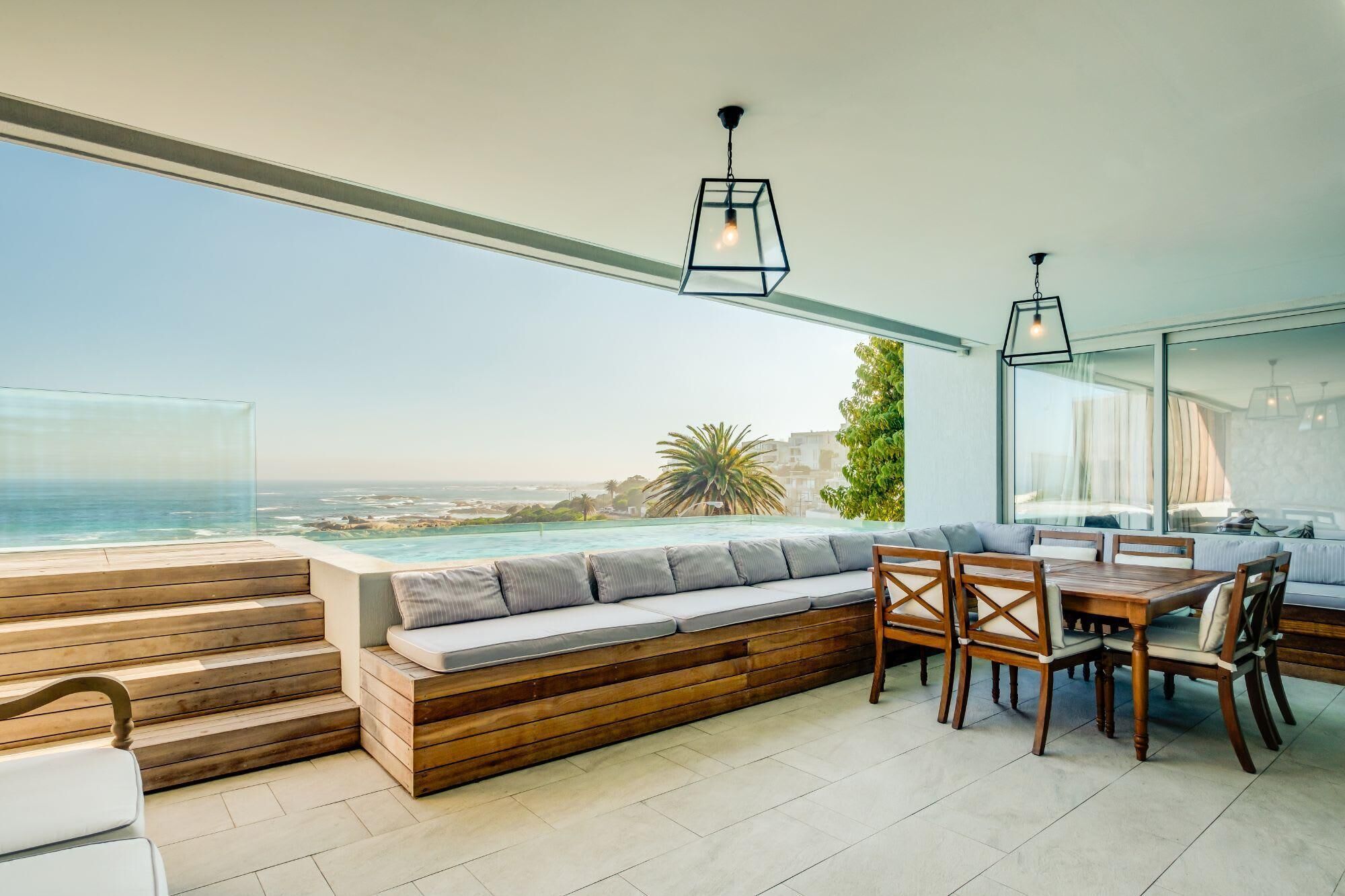 A modern outdoor seating area with a view of the ocean, wooden steps, and a dining table with chairs.