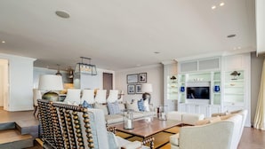 A modern living room with a large wooden chair, a sofa, and a coffee table. There is a television mounted on the wall and a bookshelf with decorative items. - Gorgeous Apartment in Secure Block w Sea Views Caliche (Cape Town)