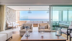 A modern living room with a large glass door leading to a balcony overlooking the ocean. There are two sofas, a coffee table, and a decorative glass candle holder. - Gorgeous Apartment in Secure Block w Sea Views Caliche (Cape Town)