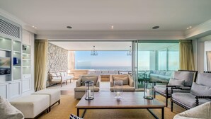 A spacious living room with a view of the ocean, furnished with sofas, a coffee table, and a TV. - Gorgeous Apartment in Secure Block w Sea Views Caliche (Cape Town)