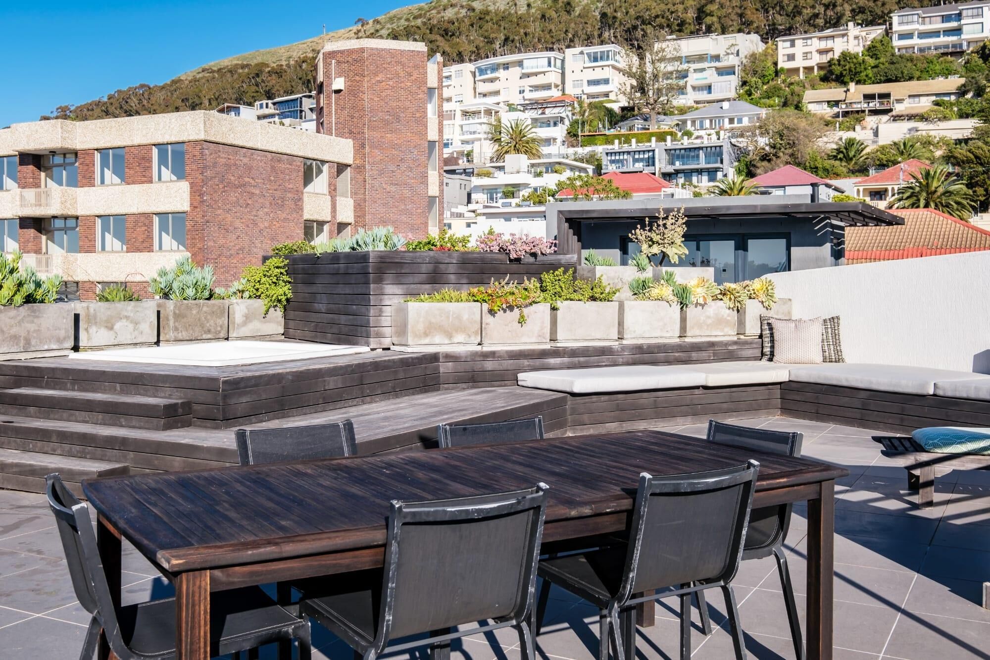 An outdoor patio with a wooden table and chairs, surrounded by planters with various plants.