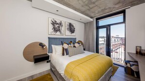 A modern bedroom with a large bed, a headboard, a bedside table, and a view of the city through a large window. - Beautifully Decorated Apartment in Cape Town Urban Signature (Cape Town)