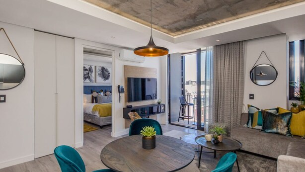 A modern living room with a round dining table, teal chairs, and a pendant light. - Beautifully Decorated Apartment in Cape Town Urban Signature (Cape Town)