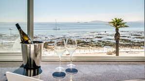 Uma garrafa de champanhe num balde de metal e duas taças sobre uma mesa, com vista para o mar e a praia.