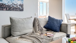 Uma sala de estar moderna com um sofá cinzento, um cobertor estampado e um travesseiro azul. Há uma revista no sofá e uma fruteira na mesa de centro.
