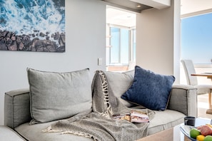 A modern living room with a grey sofa, a patterned throw blanket, and a blue pillow. A magazine is on the sofa, and a fruit bowl is on the coffee table. - Beautiful Apartment Walking Distance to the Ocean Atlantic Views (Cape Town)