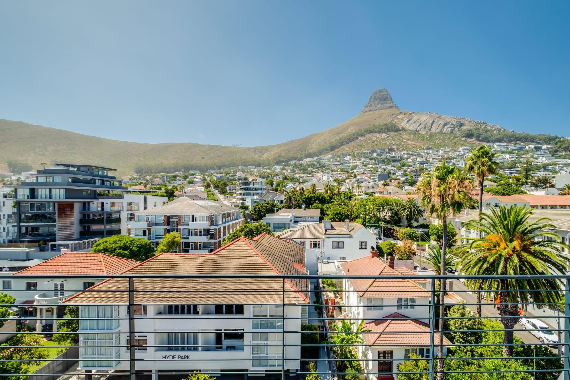 A residential area with modern houses, a prominent hill in the background, and a view through a metal fence.
