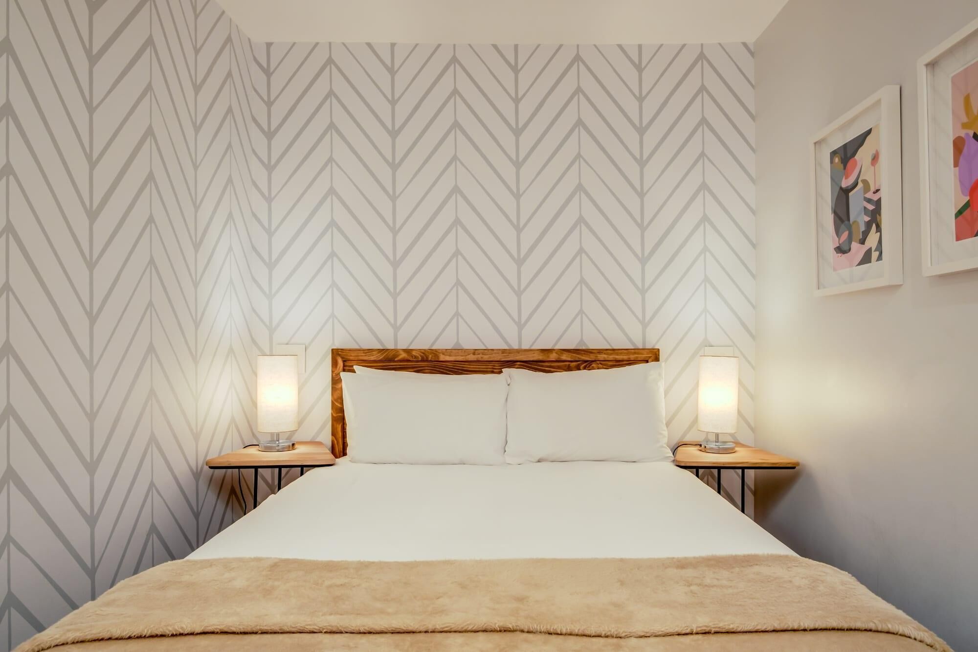 A neatly made bed with a wooden headboard, two bedside lamps, and a patterned wallpaper.