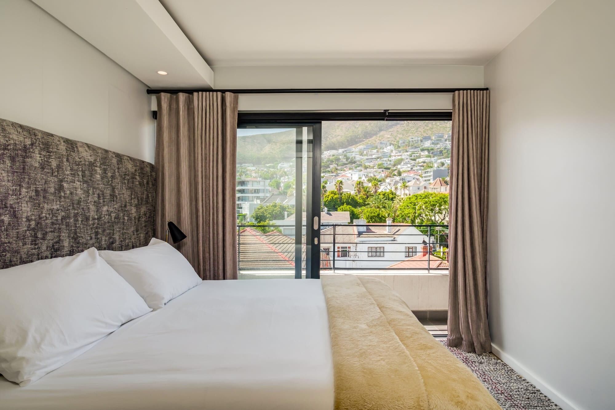 A modern hotel room with a large bed, a view of a cityscape through a sliding glass door, and a balcony with a view of the city and hills.