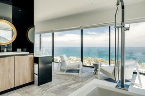 A modern bathroom with a large bathtub, a white chair, and a view of the ocean.