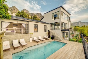 A modern two-story house with a swimming pool, lounge chairs, and a view of mountains.