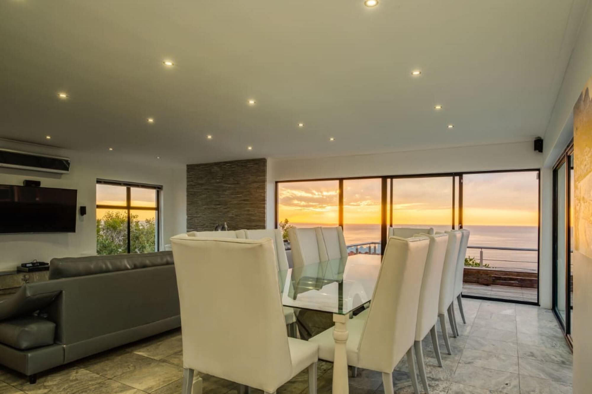 Een moderne woonkamer met een glazen eettafel en stoelen, een grijze bank en een groot raam met uitzicht op een zonsondergang.