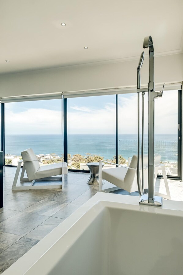 A modern bathroom with a large bathtub, two white chairs, and a view of the ocean.