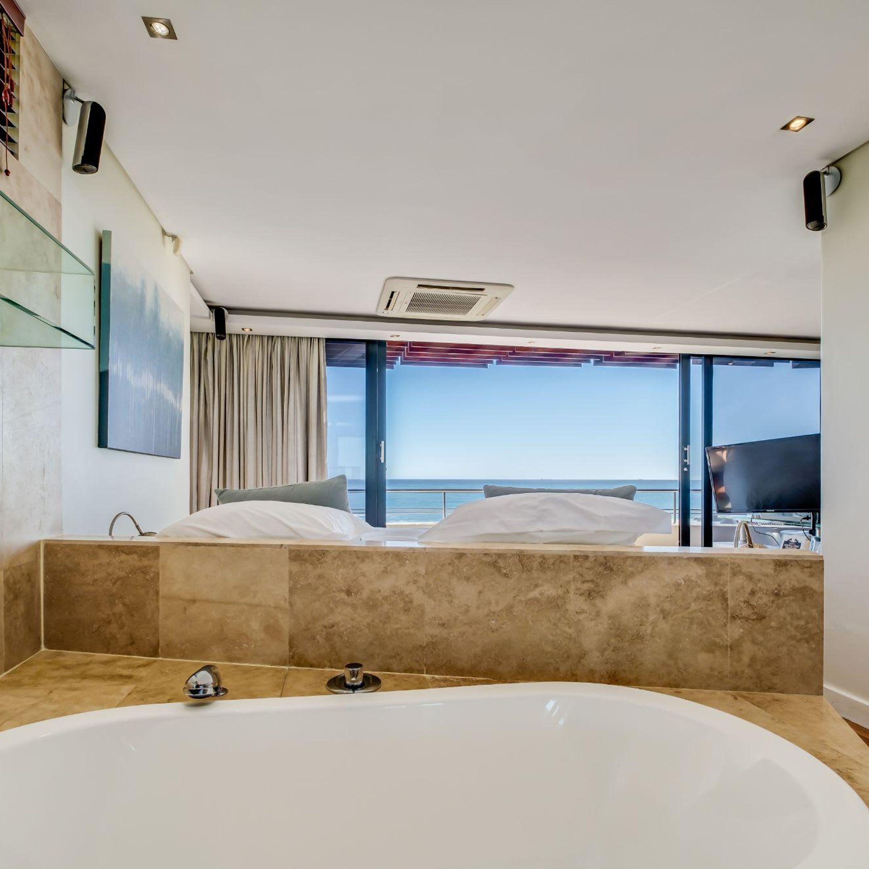 A modern hotel room with a large bathtub, a bed with white linens, and a view of the sea through large windows.