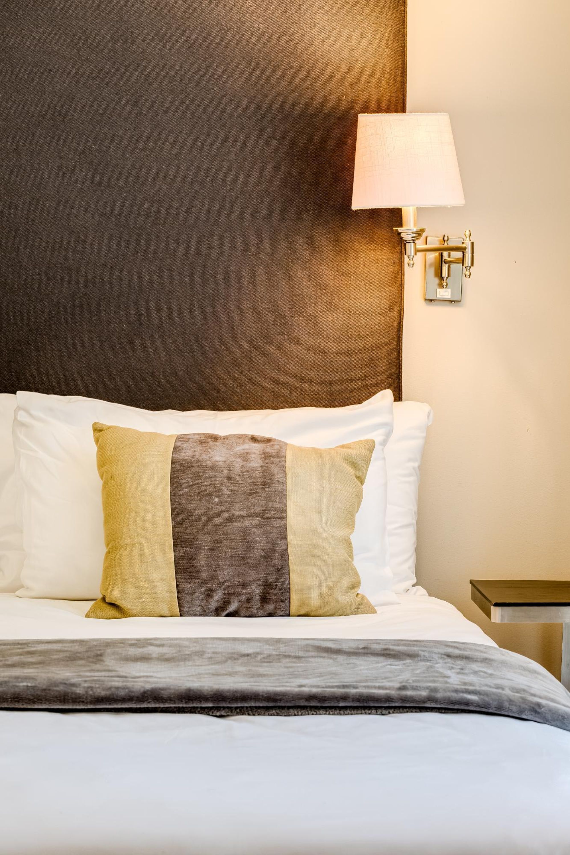 Una cama bien hecha con cabecera, una lámpara de noche y una mesita junto a la cama.