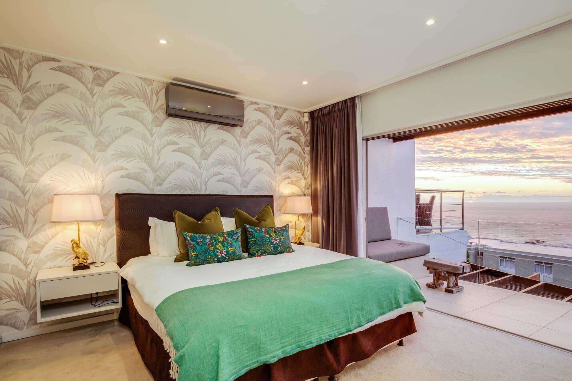 A bedroom with a bed, nightstands, a green throw blanket, decorative pillows, a view of the ocean, and a balcony with a chair.