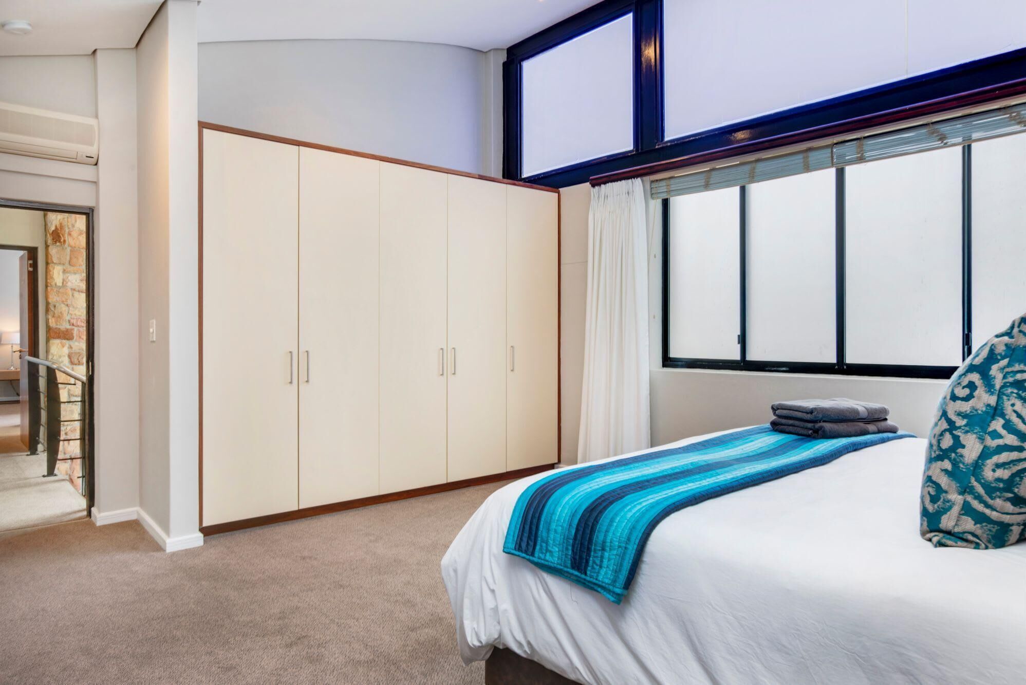 A bedroom with a bed, a large wardrobe, a window with curtains, and a view of a room with a staircase.
