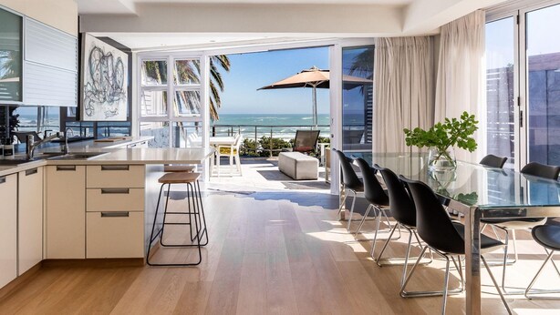 A modern kitchen with a large island, a dining area with a glass table, and a view of the ocean. - Beautiful 3bedroom Seaside Apartment Paloma Apartment (Cape Town)