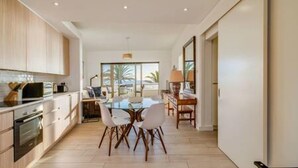 A modern kitchen with wooden cabinets, a dining area with a glass table, and a view of palm trees through the window. - Beautiful Lock up go w Ocean Views Sundowner Views (Cape Town)