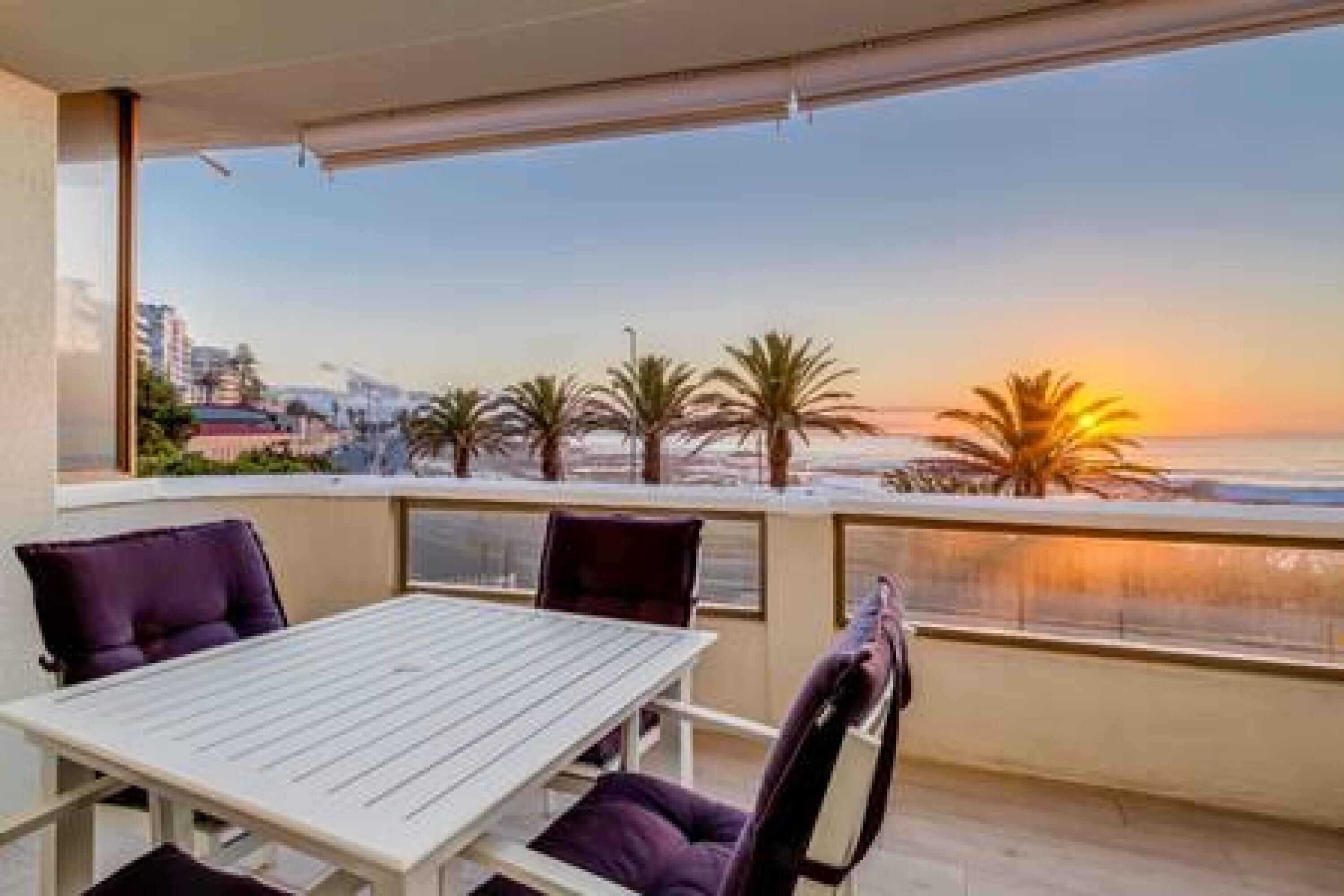 A balcony with a table and chairs, overlooking a beach with palm trees and a sunset.