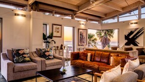 A spacious living room with a large brown leather sofa, two grey armchairs, and a coffee table. There are several framed artworks on the walls, a large window with a view of palm trees, and a wooden ceiling structure. - Unique Villa w Pool Garden and Sea Views Lobster Villa (Cape Town)