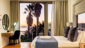 A bedroom with a bed, a desk, a chair, and a view of the sunset and palm trees. - Unique Villa w Pool Garden and Sea Views Lobster Villa (Cape Town)