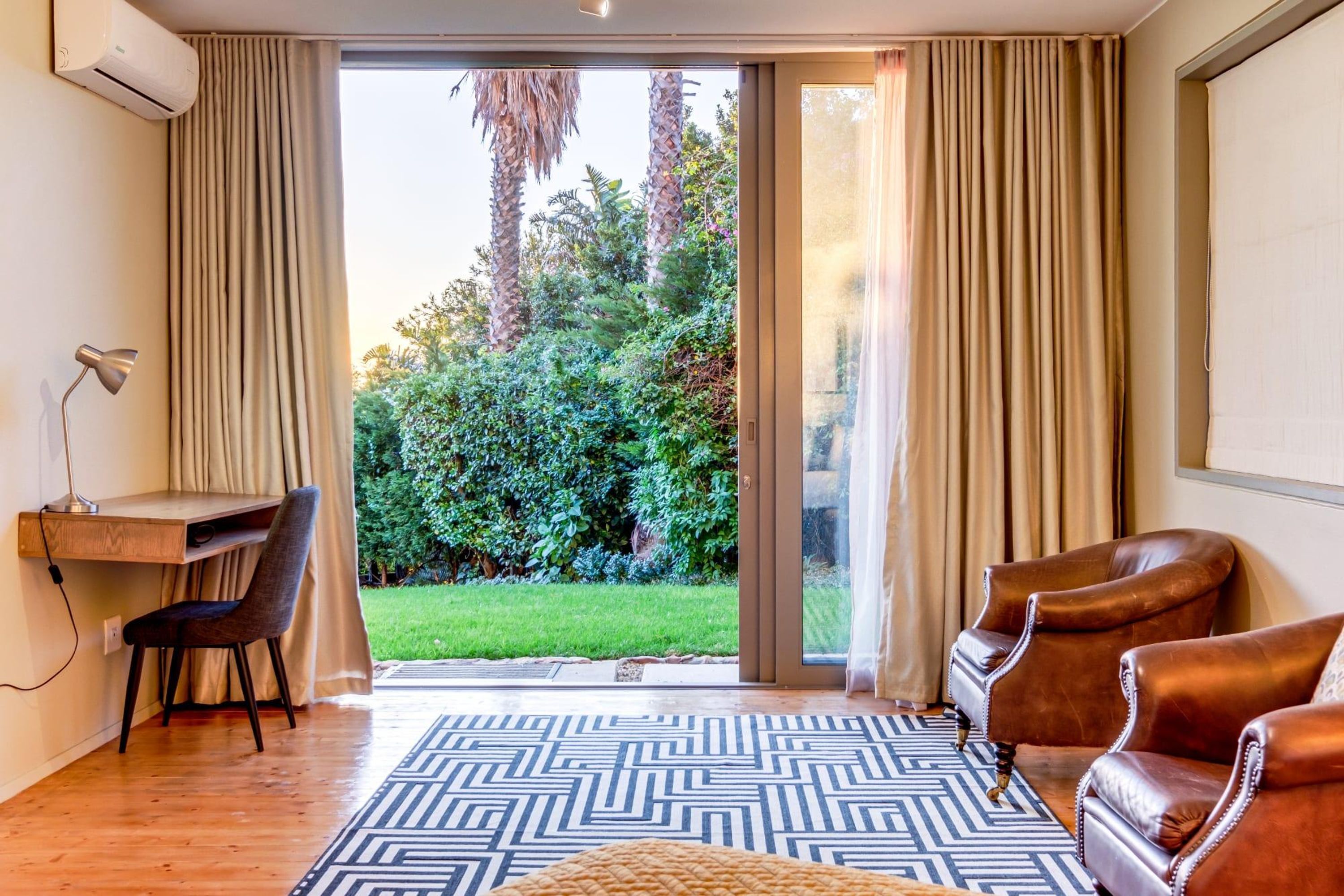 A room with a desk, chair, two leather armchairs, a patterned rug, and a sliding glass door leading to a garden with trees and grass.