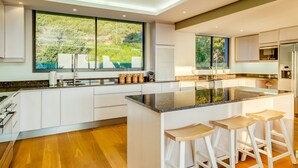 Une cuisine moderne dotée d’un grand îlot central, de meubles blancs et offrant une vue sur la verdure environnante.