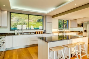 A modern kitchen with a large island, white cabinetry, and a view of greenery outside.
