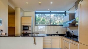 A modern kitchen with a central island, built-in oven, and a large window providing a view of trees. - 308 Juliette on the Waterfront Marina (Cape Town)