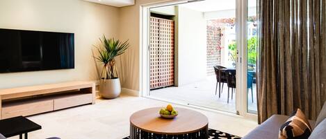 A modern living room with a flat-screen TV, a round wooden coffee table with fruit, a sofa, and a sliding glass door leading to an outdoor area.