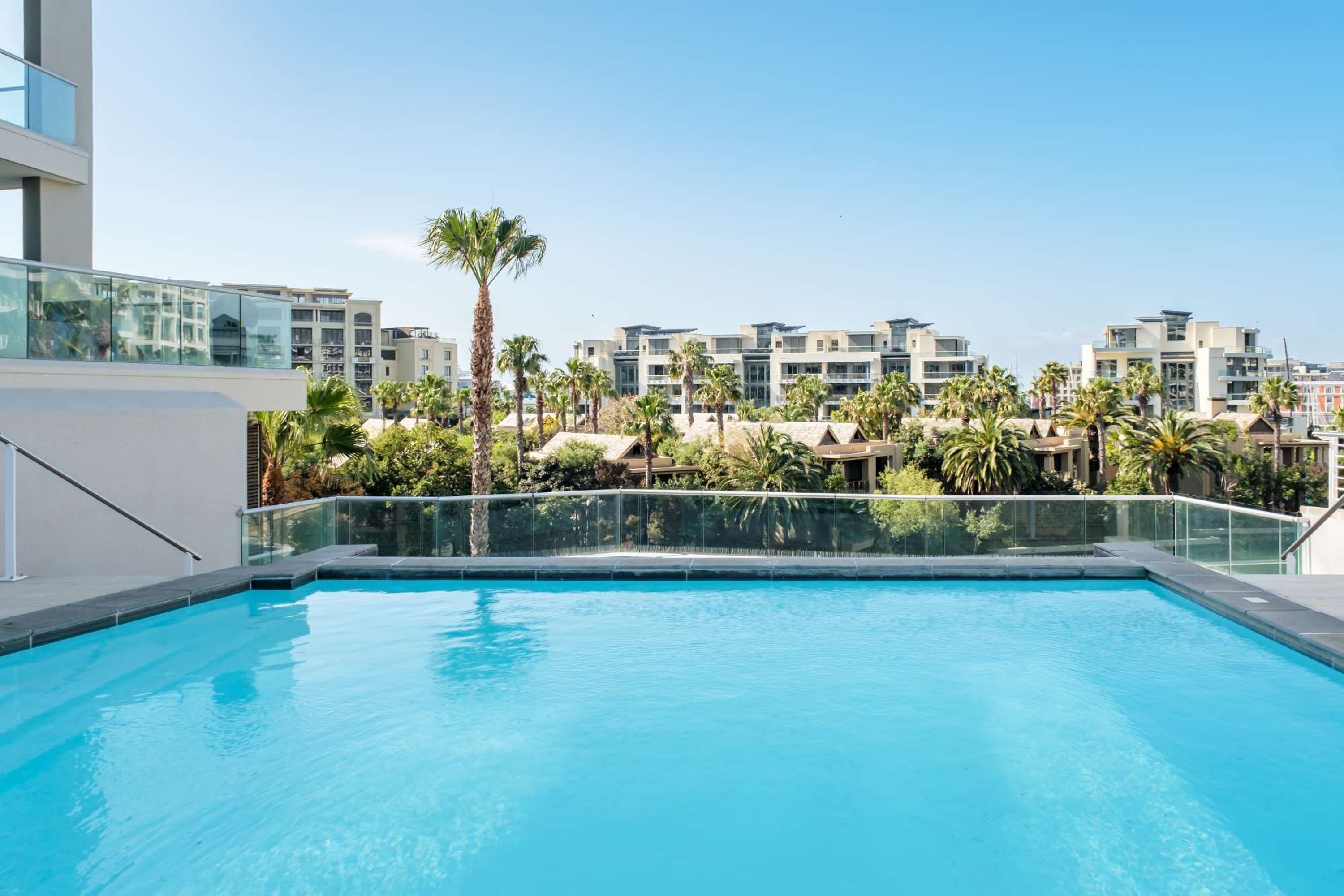 A swimming pool with a glass railing, surrounded by palm trees and modern buildings.