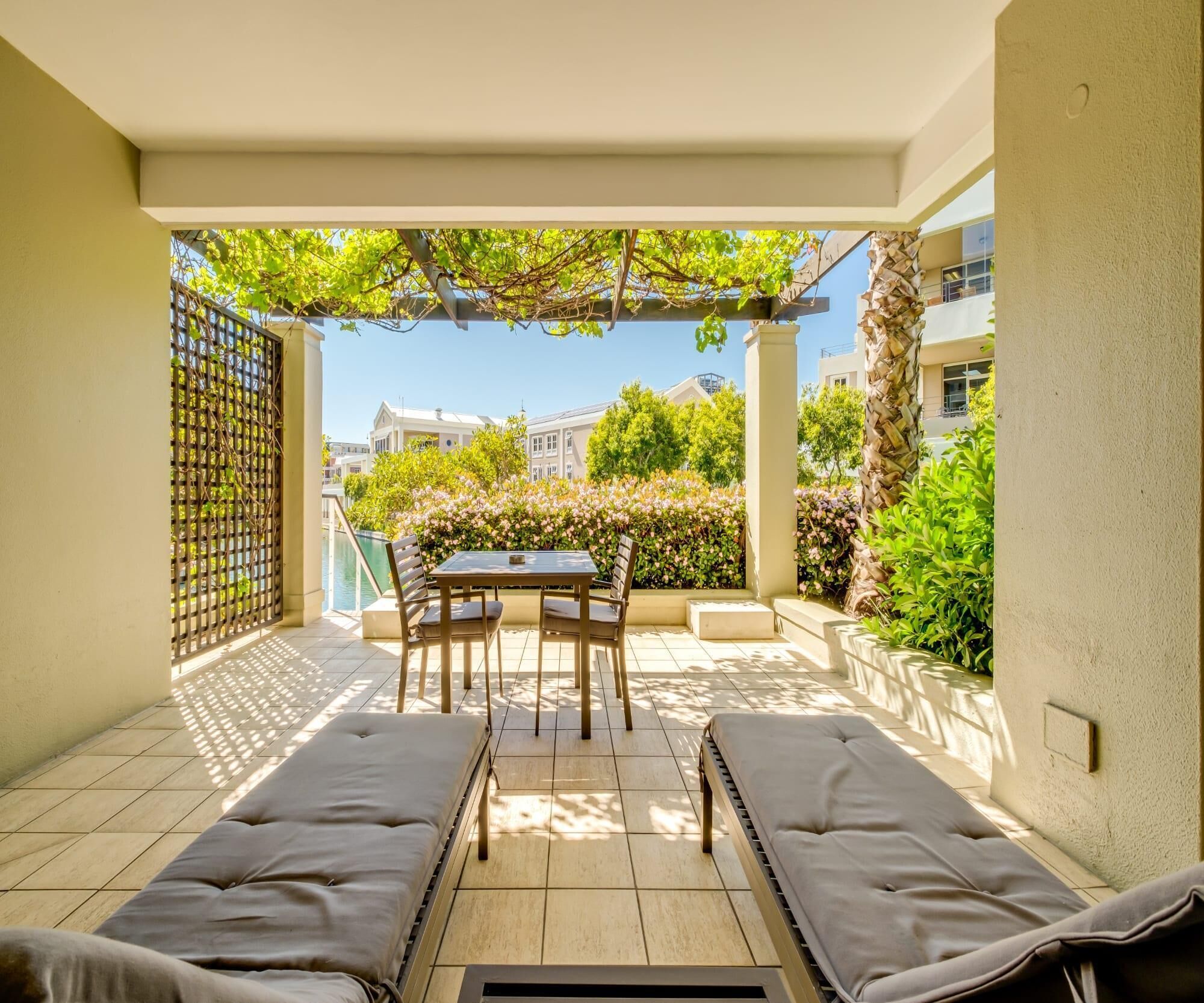 A sunlit patio with a table and chairs, two cushioned benches, and a view of a pool and buildings in the background.