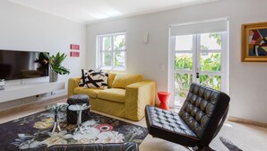 A living room with a sofa, a black leather chair, a flat-screen TV, a coffee table, and a large window with a view of greenery. - Unique City View Apartment in De Waterkant 207 DWP (Cape Town)