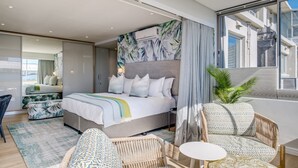 Quarto de hotel moderno com cama grande, pequeno sofá, mesa de jantar e uma janela ampla com vista para a cidade.
