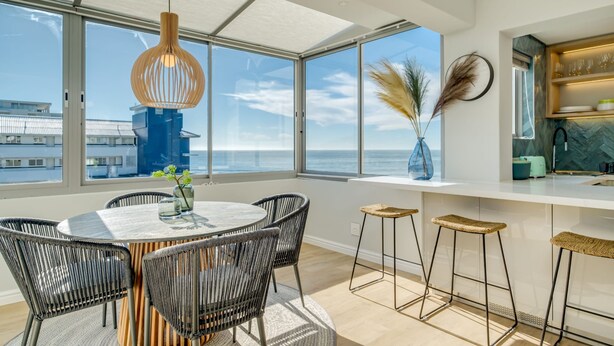 Uma cozinha moderna com bancada branca, banquetas e vista para o oceano.