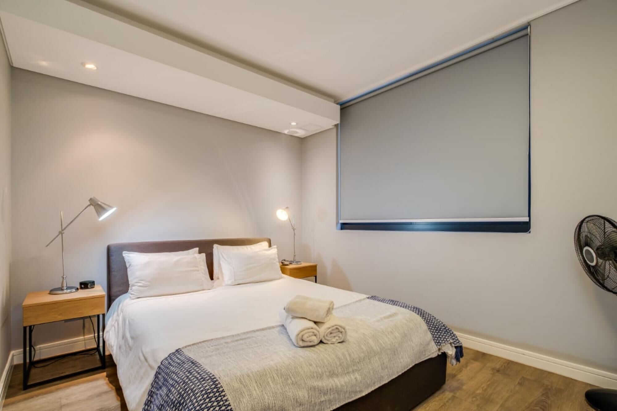 Quarto com cama, abajures, ventoinha na parede e uma janela grande.