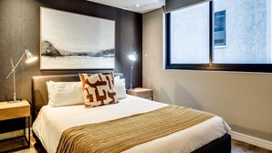 Un dormitorio moderno con una cama grande, una mesita de noche con lámpara, un mueble auxiliar junto a la cama y una ventana que ofrece vistas a un edificio, además de un cuadro abstracto de gran tamaño en la pared.