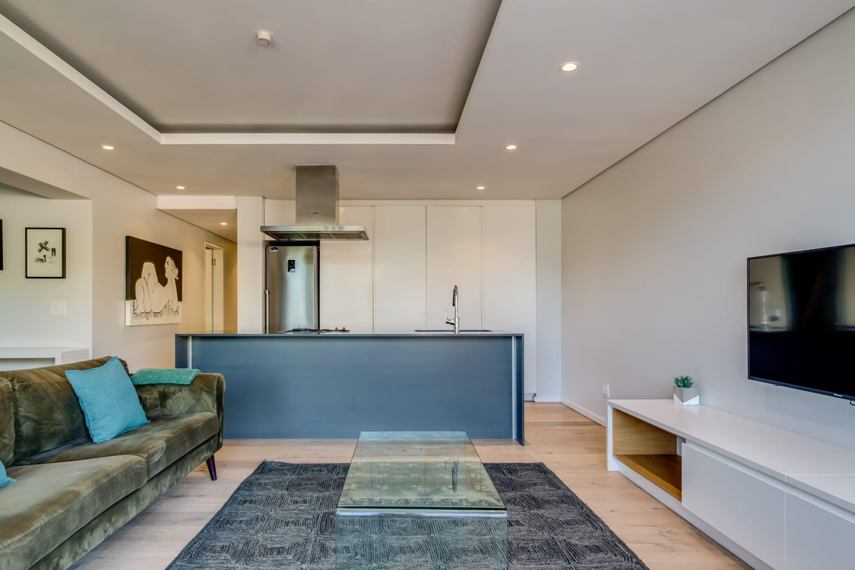 A modern living room with a sofa, a glass coffee table, a flat-screen TV, and a kitchen area with a built-in island.