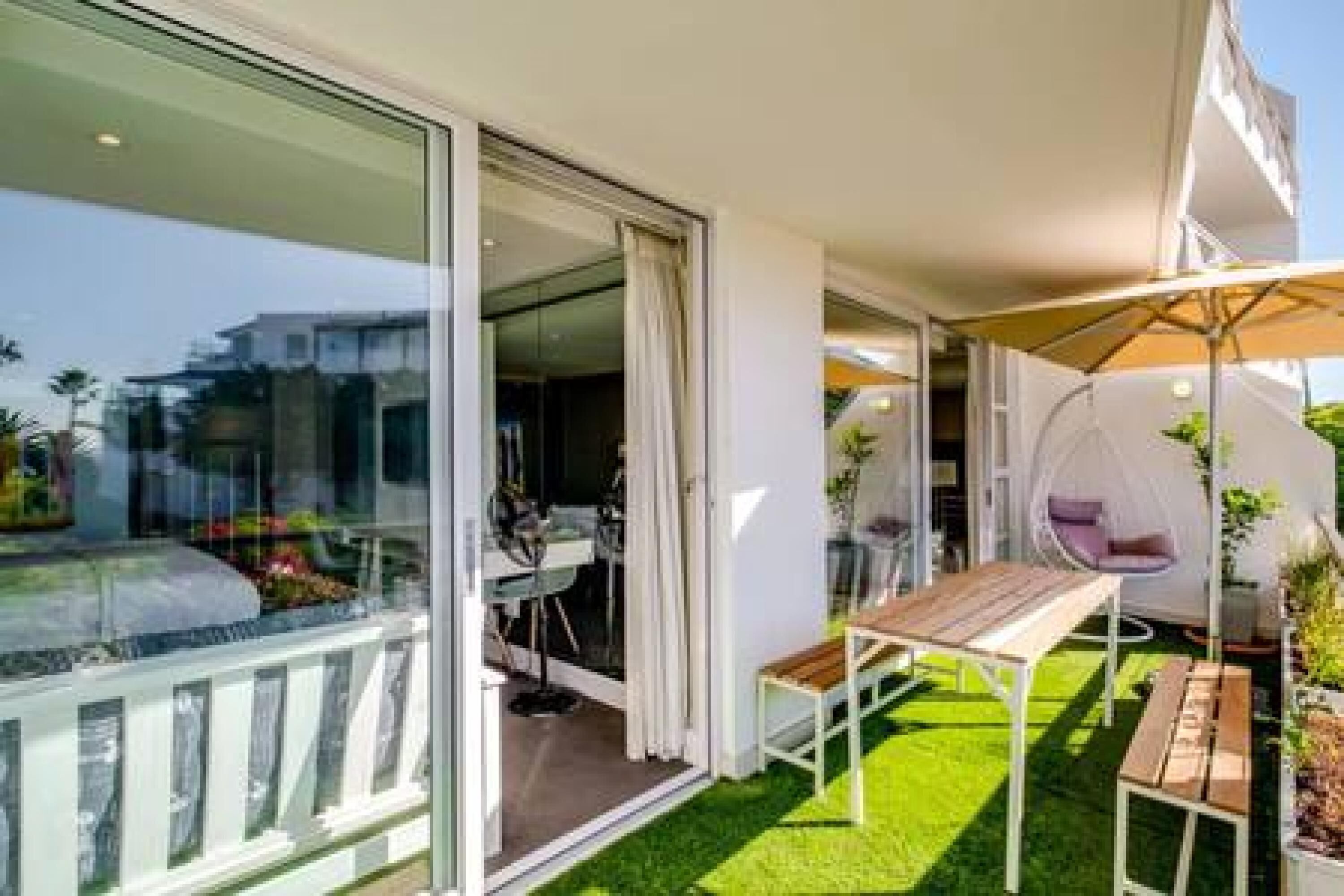 A modern outdoor patio with a glass door leading to a room, a wooden table, a cushioned chair, and a hanging chair.