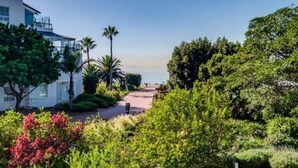 A pathway leading to a beach, surrounded by lush greenery and a modern building with large windows. - Elegant Apartment at the Waterclub Amani Views (Cape Town)