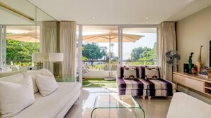 A modern living room with a sofa, a coffee table, and a view of a garden through large windows. - Elegant Apartment at the Waterclub Amani Views (Cape Town)