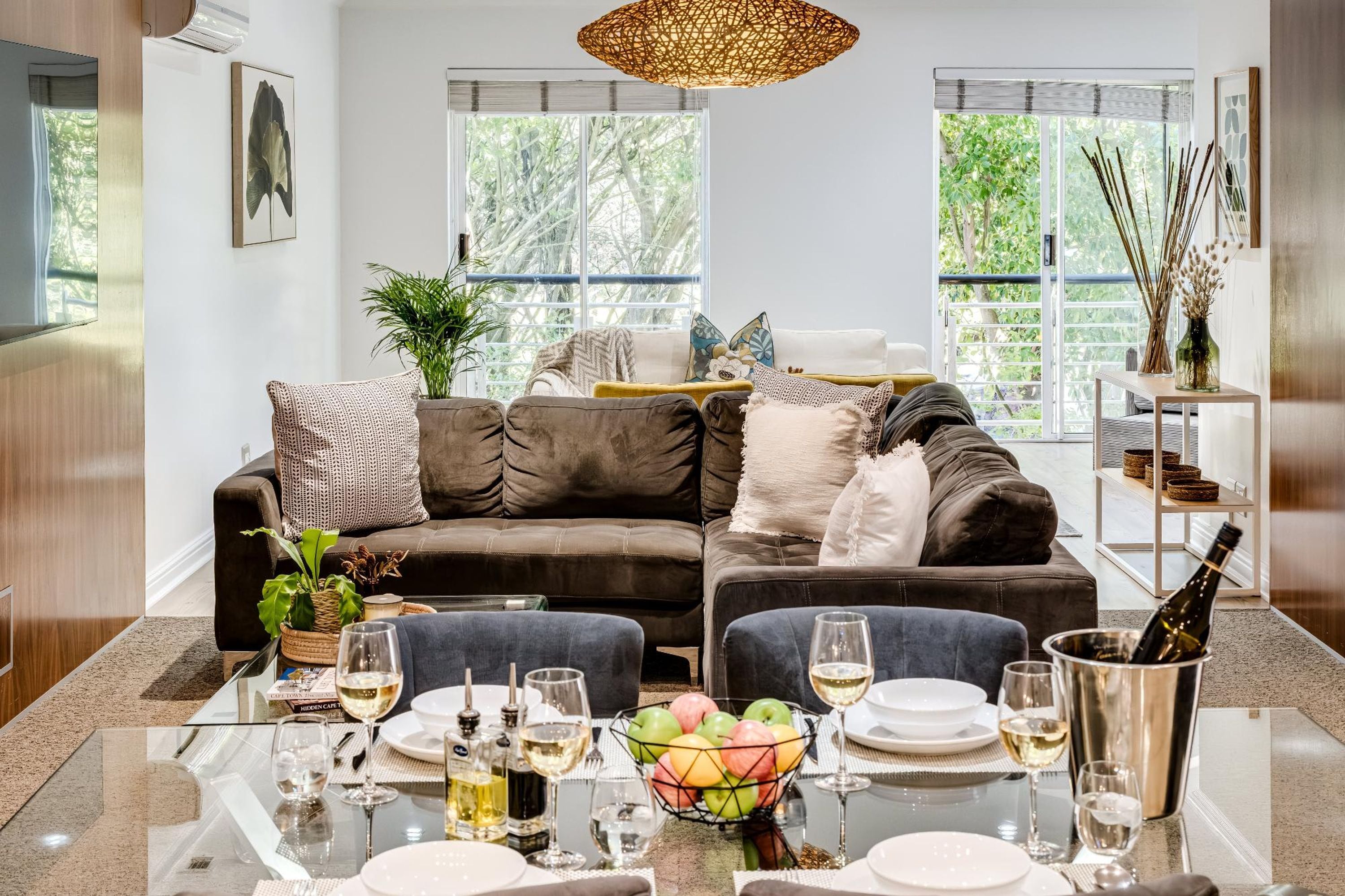 A modern living room with a sectional sofa, a dining table set for a meal, and large windows offering a view of greenery.