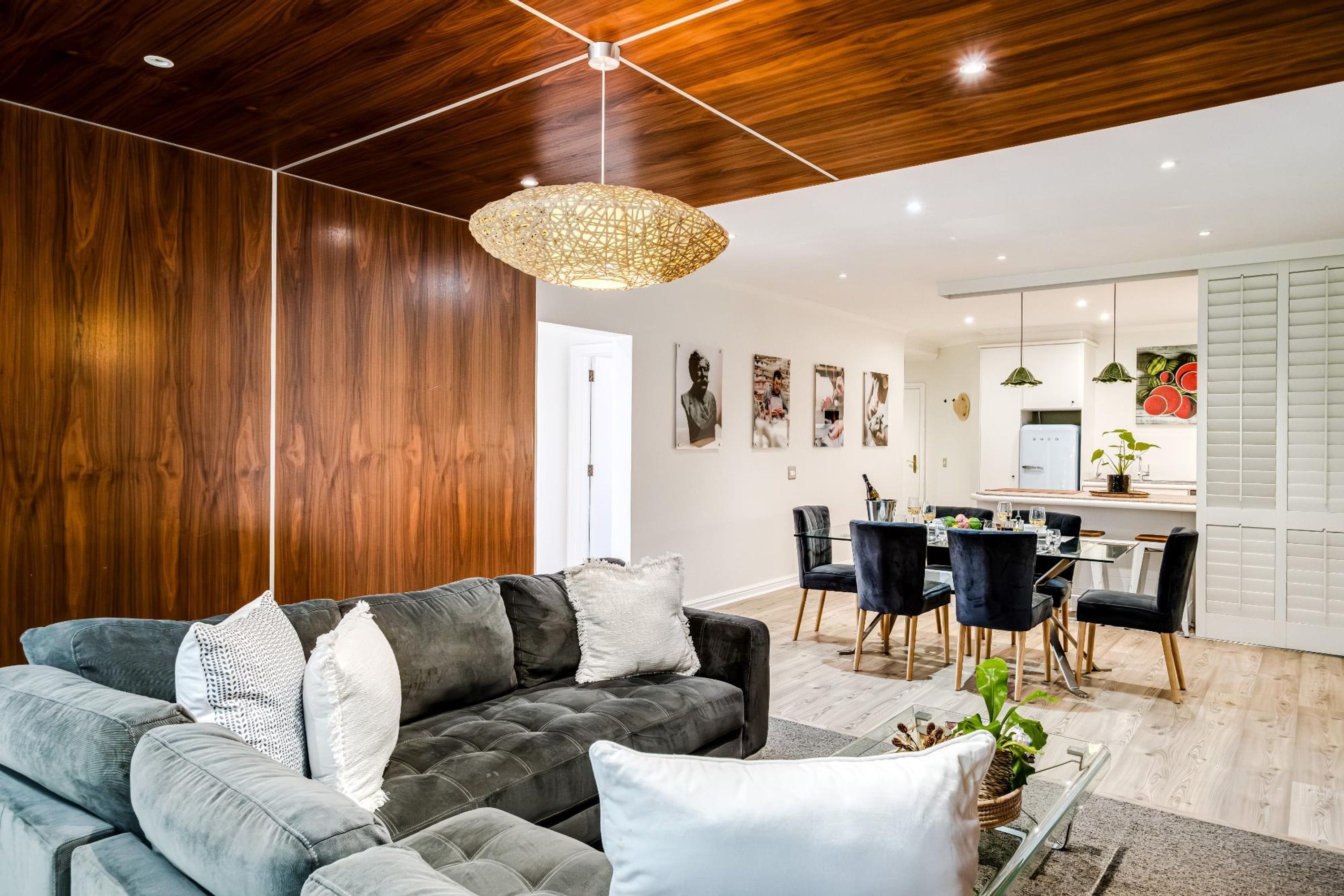 A modern living room with a dark grey sofa, white and grey cushions, a wooden ceiling, and a dining area with a white table and chairs.