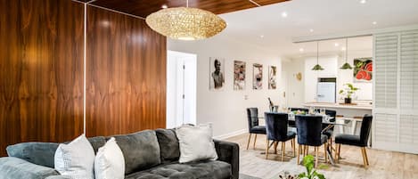 Una sala de estar moderna con un sofá gris oscuro, cojines blancos y grises, techo de madera y una zona de comedor con una mesa y sillas blancas.