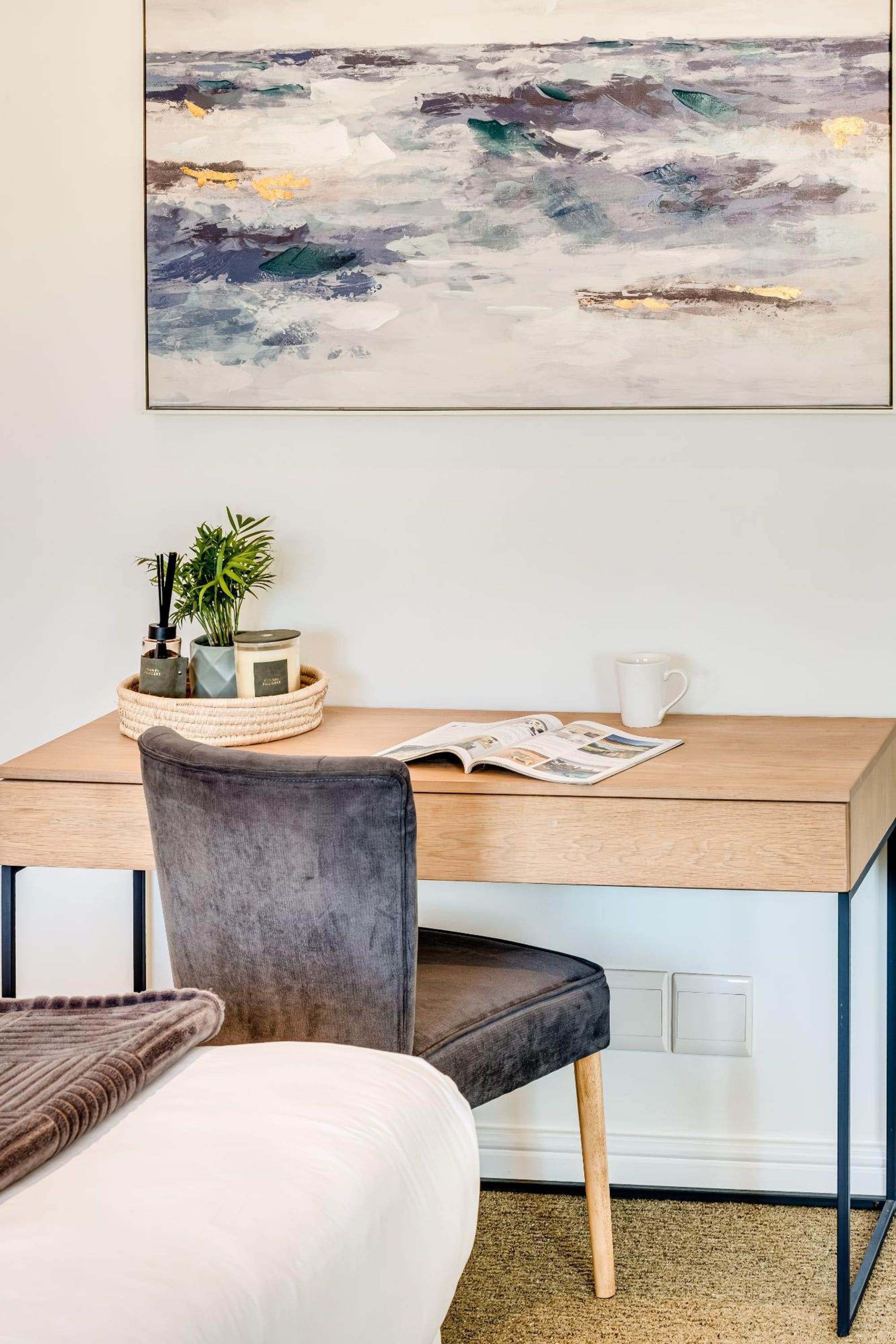 Una habitación moderna con un escritorio de madera, una silla gris, una cama con edredón blanco, una planta pequeña, una vela y un cuadro abstracto enmarcado en la pared.