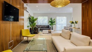 Un salon moderne avec un téléviseur à écran plat, des murs en bois, une table basse en verre, un canapé beige et un fauteuil jaune.