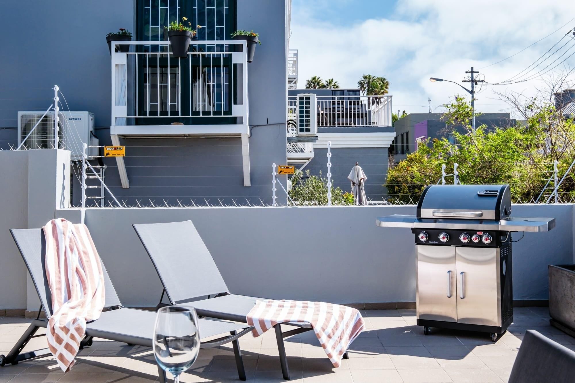 A rooftop terrace with two lounge chairs, a glass of water, a barbecue grill, and a small cabinet.